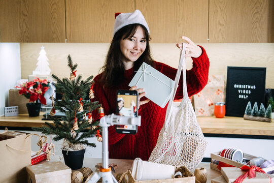 Woman in Santa hat shows gift box and mesh bag to phone on tripod near CHRISTMAS VIBES ONLY sign. Eco friendly christmas gift review, sustainable gift idea video, reusable bag gift presentation