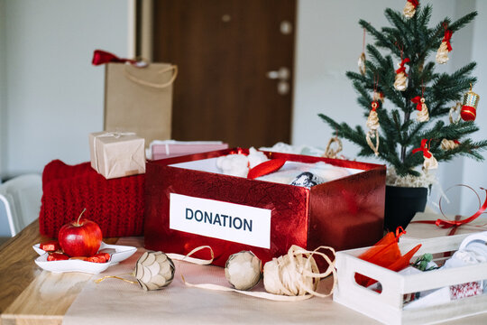 DONATION box packed with knitwear, toy and heart stands among wrapped gifts near Christmas tree. Christmas donation box tree, charity clothing box holiday, giving back christmas season
