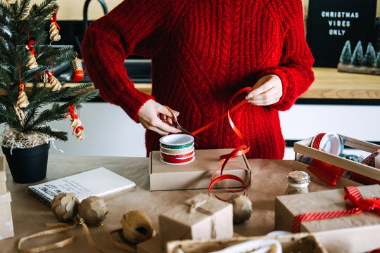 Woman pulls ribbon across kraft box on kitchen table beside decorated tree and wrapping supplies. Handmade versus mass produced, artisanal craft movement christmas, slow made gifts concept