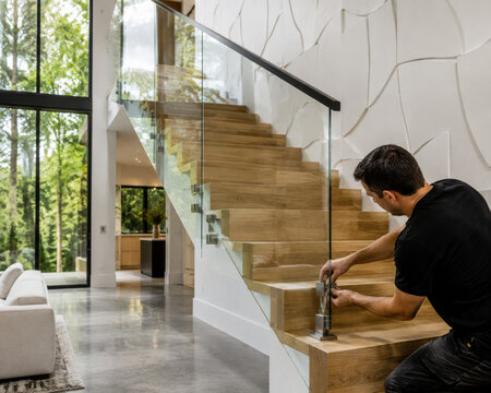 Un hombre trabaja en la instalaci&oacute;n de una escalera de madera con pasamanos de vidrio en una casa moderna rodeada de &aacute;rboles