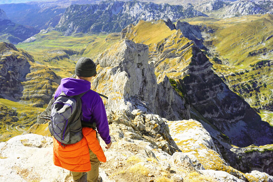 Traveler gazing from Bobotov Kuk peak at karst valley with isolated limestone spire, golden slopes, and distant summits. Depicts contemplative mountaintop pause and grandeur of Montenegrin highlands.