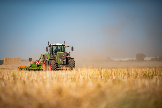 A Fendt tractor pulls a cultivator across a golden stubble field with stacked straw bales in the background, kicking up dust under a clear blue sky in the Eure region, France.