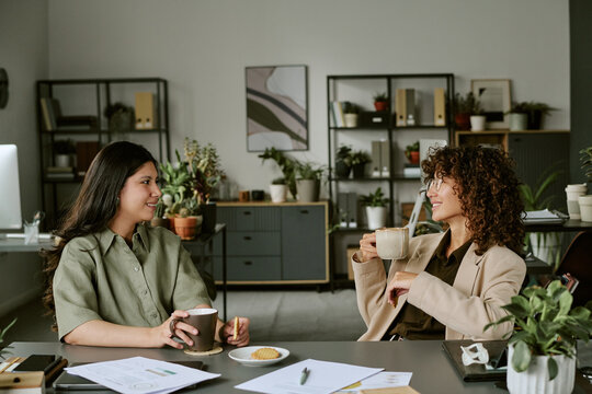 Diverse young adult female colleagues talking during coffee break in modern office, representing corporate communication and positive workplace culture, medium shot