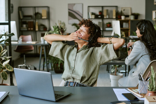 Tired young adult diverse women wearing headsets working in call center, yawning and stretching at desk. Image suitable for human resources, burnout, and mental wellbeing campaigns