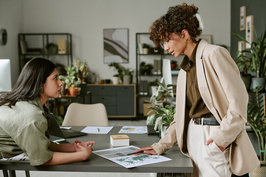 Diverse young adult female colleagues discussing financial charts at desk in modern office, representing teamwork and corporate communication, medium shot