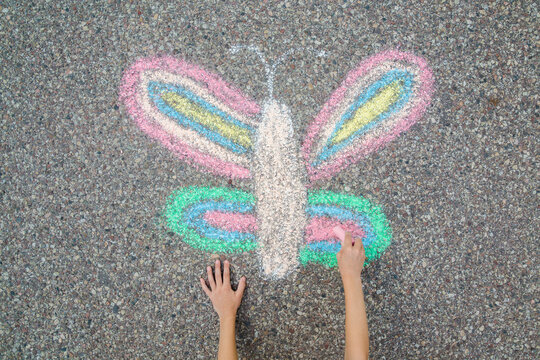 Little child girl hand fingers holding chalk and drawing colorful butterfly on dark gray asphalt background in summer day. Closeup. Point of view shot. Top down view.