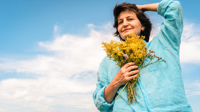 Happy woman holding bouquet of wild yellow flowers, enjoying freedom and summer under a clear blue sky