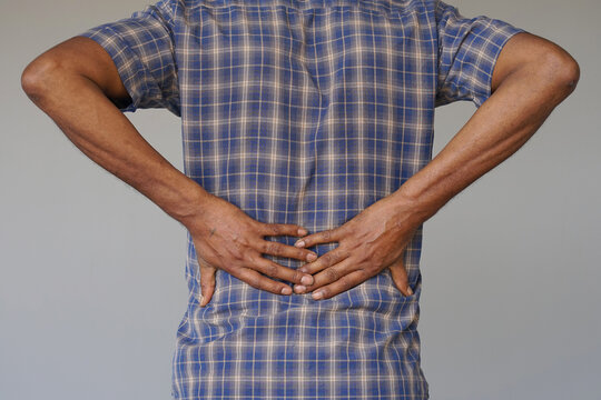 Blac man grips his lower back, showing discomfort. He wears a blue plaid shirt, standing still. His posture suggests pain or stiffness in the spine