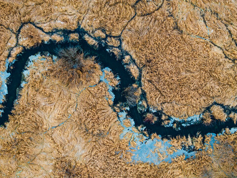 Overhead top-down aerial drone view of a complex winding river with fractured ice networks carving through a vast, highly textured brown winter wetland. Abstract natural landscape photography.