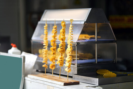 Golden fried spiral potato chips skewered on long wooden sticks displayed at street food vendor stall. Crispy spiral twisted potato skewers displayed on wooden stand at street food market counter