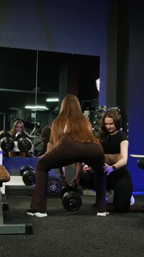 Sportswoman strengthens legs through controlled dumbbell squats. Attentive coach carefully explains important technical details. Vertical video