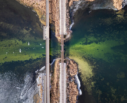 Overhead top-down aerial drone view of two parallel bridges crossing a channel of vibrant green water flanked by rocky banks. Geometric abstract infrastructure and nature photography.