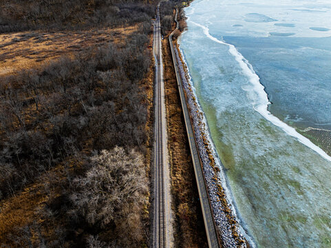High-angle aerial drone view of straight railroad tracks running parallel to a frozen lake shoreline and a bare winter forest. Scenic seasonal infrastructure landscape photography.