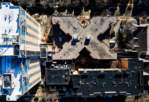Overhead aerial drone view of a distinct X-shaped urban building structure nestled between high-rise apartments and city blocks. Conceptual top-down architectural cityscape photography.