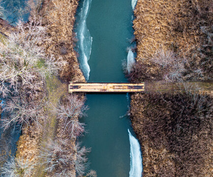 Aerial view of a narrow bridge crossing a calm blue river surrounded by brown vegetation and patches of ice, capturing the serene geometry and seasonal contrast of late autumn or early winter.