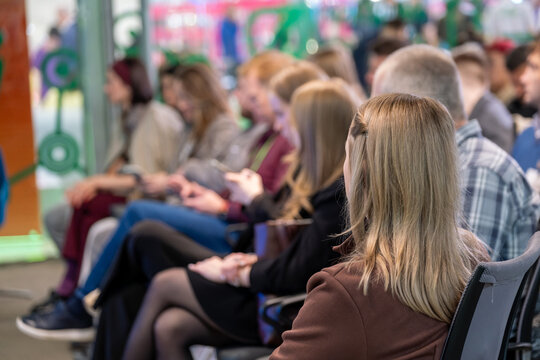 Woman sits among conference attendees and listens from a row of chairs in a softly blurred event space.