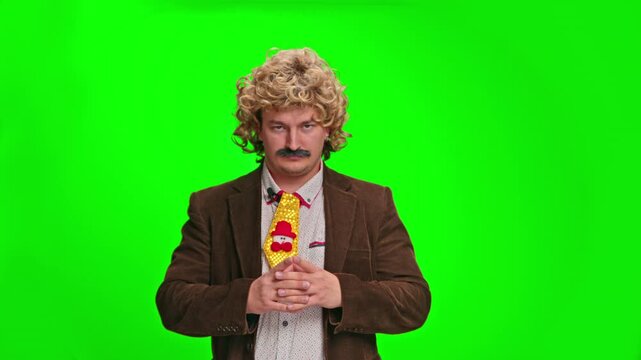 An adult man wearing a curly blonde wig, fake mustache and brown blazer turns from facing away to facing forward, gesturing with his hands against a green screen background.