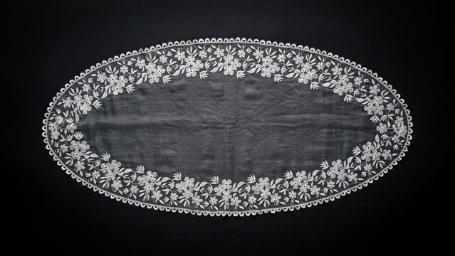 Vintage white lace doily with floral embroidery. Antique oval table runner on black background. Elegant handmade textile craft.