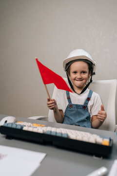 Happy child in denim overalls and construction helmet plays at office desk. She holding red flag, smiling showing thumbs up, symbolizing future ambition and success.