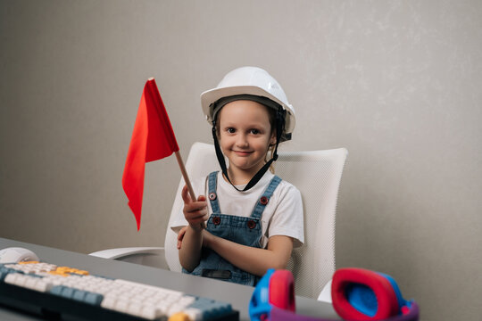 Young child wearing white safety helmet and blue overalls, smiling looking at camera while holding red warning flag at desk with keyboard, symbolizing safety and future careers.