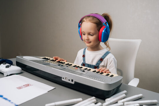 Little girl concentrating on digital keyboard, wearing colorful headphones and smiling while learning music at home, developing creative skills and enjoying playful musical hobby.
