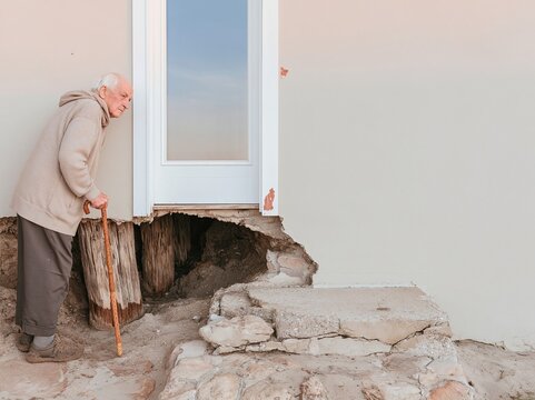 Senior man with a walking cane inspecting an undercut foundation after coastal erosion for insurance claims and house repair services