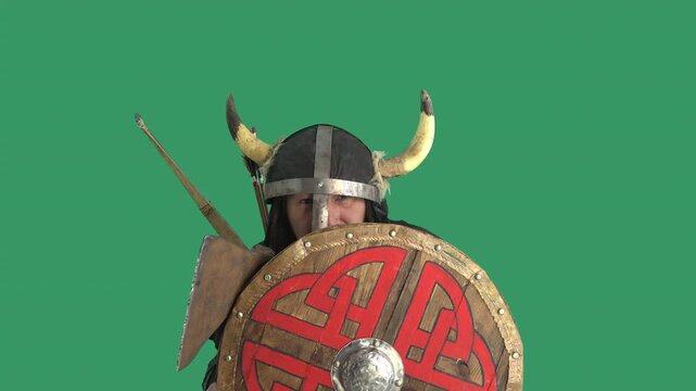 Viking warrior in horned helmet hides behind a round wooden shield from arrows and nervously adjusts the helmet with his axe, isolated on a green screen for chroma key