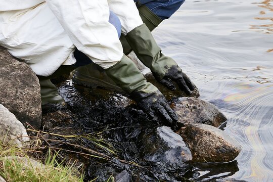 Oil spill response worker in protective clothing manually cleaning petroleum-covered rocks along a contaminated shoreline