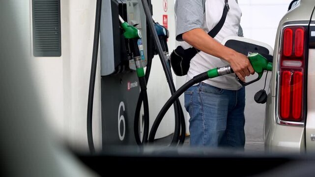 Man fills car at gas station. Holding nozzle, he concentrates. Vehicle's tail light glows red. Frame shows gas pump, numbered 6. Daily routine, simple, unembellished. Use for automotive, travel