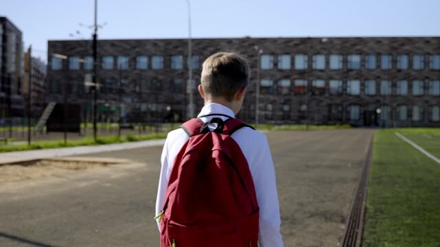 Boy walks toward school, red backpack visible. Bright day, clear sky, brick building in background. Schoolyard grass, asphalt path, distant goalpost. Quiet moment, no facial expressions