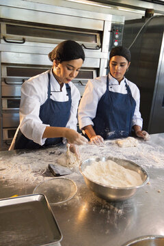 Diverse female bakers wearing jackets and hairnets shaping dough at bakery kitchen near deck oven