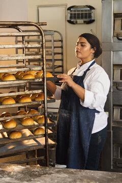 Woman in chef coat arranging trays of golden rolls on metal rack at bakery, copy space
