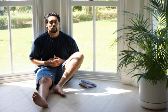 Man sitting barefoot on wood floor by multi-pane window with journal, pen and palm