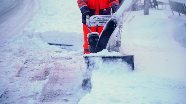 Worker clears snow with mechanical snow blower. Snow flies as machine grinds thick frost from road. Red suit contrasts snowy landscape. Utility worker performs essential winter task