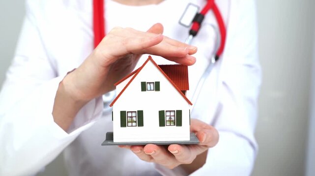 Doctor gently shields a miniature house. Hands show care and protection. White coat and stethoscope suggest professionalism. Ideal for insurance or home safety concepts