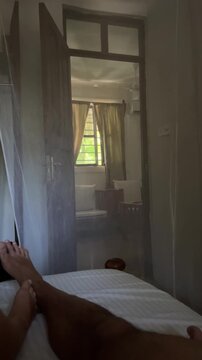 Vertical HD POV video of a person lying in bed under a canopy mosquito net in a tropical resort room in Zanzibar. Looking towards the open door leading to a sunlit living space.