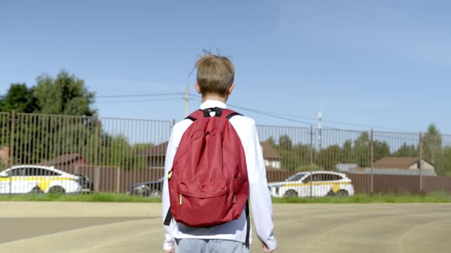 Young boy walks away from school with red backpack. Bright sky and clear weather surround him. School gates and parked cars line the road. He appears calm and focused on his path