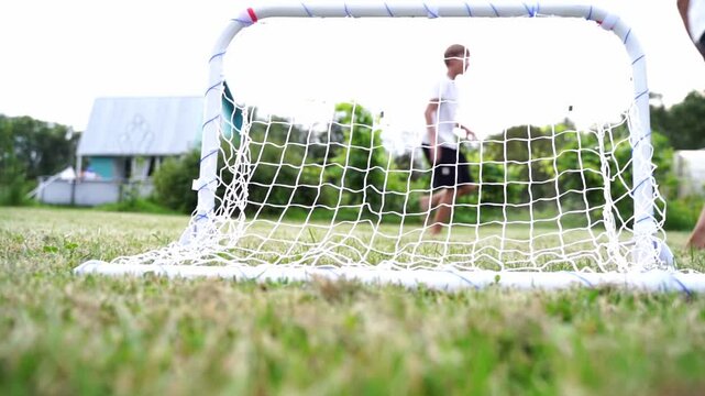 Children play soccer near small goal on grassy field. One child runs toward goal, another stands nearby. Sunlight casts soft shadows on green grass. Game feels energetic, carefree, and fun