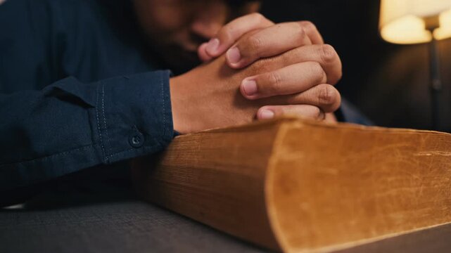 Christian man holding clasped hands together on old bible book while praying in warm dim light room, seeking guidance, wisdom, peace, spirituality, study gospel and worshipping Lord God.