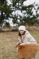 Naklejka premium Girl in a warm beige coat and white beanie outdoors, smiling as she pats a fluffy alpaca in a grassy field on a cool day, sharing a wholesome nature moment