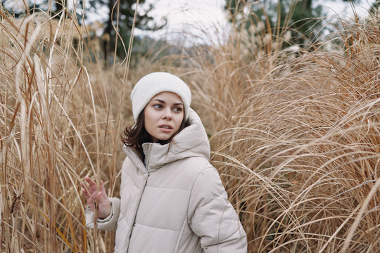 Winter scene with a woman in a puffy beige coat and white beanie standing among tall dry grass, calm expression, outdoor portrait against a natural rustic field and soft overcast light.