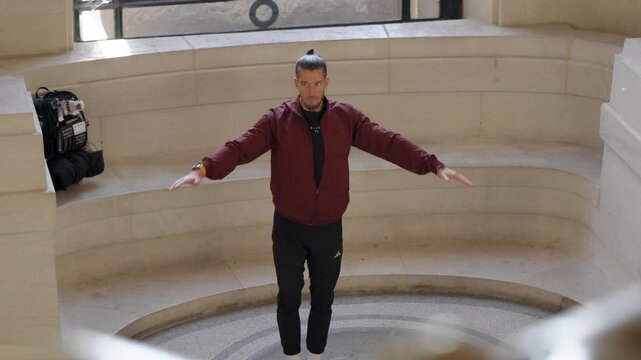 High-angle shot of a male athlete in a burgundy jacket stretching his arms out in a stone circular hall before an outdoor workout session.