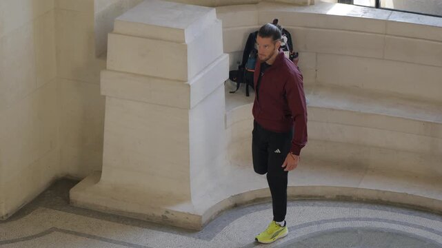 High-angle shot of a male athlete in a burgundy jacket stretching his quadriceps in a stone circular hall before an outdoor workout.