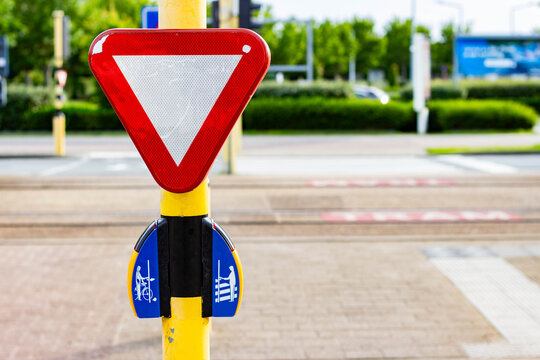 Traffic sign indicating yield with bicycle and pedestrian symbols mounted on a yellow pole near a crosswalk in an urban environment with greenery in the background