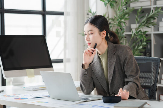 Focused Professional at Desk: A focused young woman in professional attire works diligently at her desk, surrounded by office supplies, a computer, and paperwork, in a bright and modern workspace.