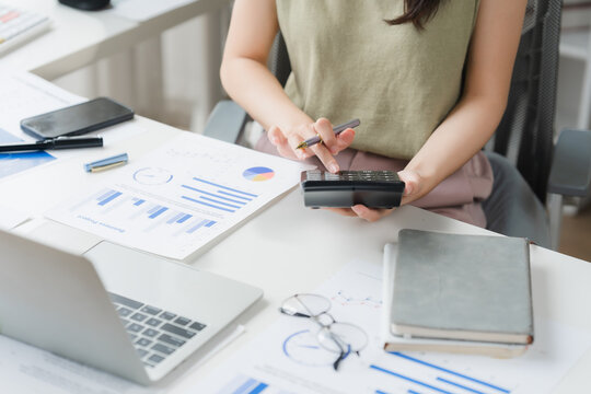 Business Woman Calculating Profits: A focused business woman sits at her desk, meticulously calculating figures on a calculator surrounded by financial reports, a laptop, and glasses.