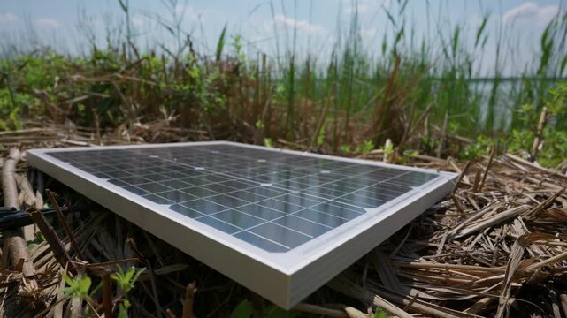 A close-up view shows a compact portable solar panel resting on dry grass near a lake. The high quality photovoltaic equipment collects sunlight to generate clean green energy outdoors.