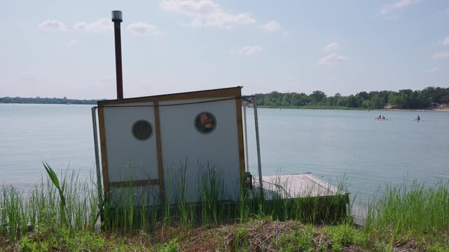 A homemade floating sauna cabin rests along a grassy lake shoreline in bright daylight. The small wooden structure features a chimney pipe and circular windows for summer leisure.
