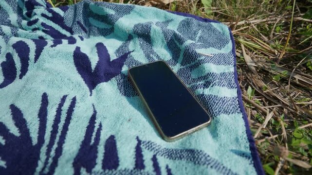 A dark smartphone rests on a blue patterned beach towel laid on dry grass in the sun. Concept of digital detox, summer vacation, outdoor relaxation, and modern connectivity.