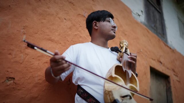 Young man in white shirt plays a carved sarangi with bow while standing beside a weathered orange mud wall of a rural Himalayan house